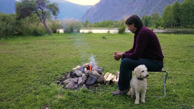 family vacation destination on the banks of the river in the mountains around the campfire. A middle-aged Caucasian man in a bard sweater playing with a white dog breed poodle. evening light of a summ