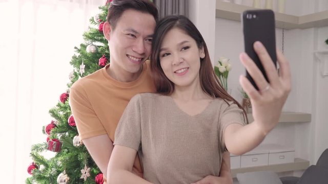 Asian couple using smartphone selfie with christmas tree decorate their living room at home in Christmas Festival. Lifestyle woman and man happy celebrate summer christmas and New year concept.