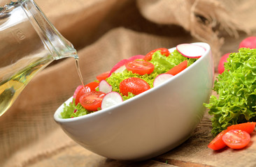 Pouring oil into green salad Lollo Biondo with tomatoes and radishes in a white bowl on wooden table, a pitcher of oil in the background