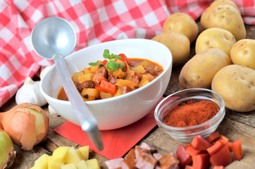 Potato and sausage goulash in dish, pepper, garlic, onion and ladle on wooden table