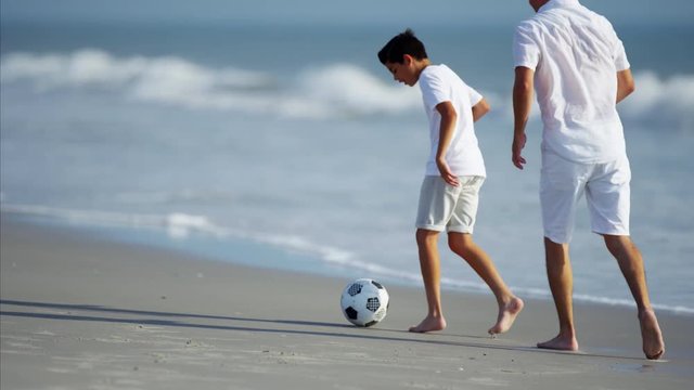 Latin American father and son playing with soccer ball on beach holiday