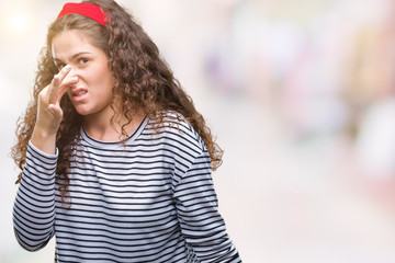 Beautiful brunette curly hair young girl wearing stripes sweater over isolated background smelling something stinky and disgusting, intolerable smell, holding breath with fingers on nose