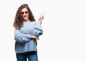 Beautiful brunette curly hair young girl wearing sunglasses over isolated background smiling with happy face winking at the camera doing victory sign. Number two.