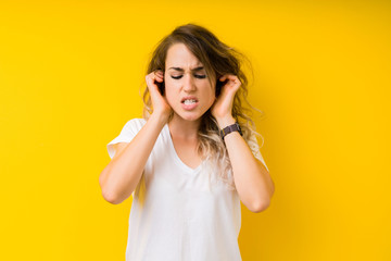 Young beautiful blonde woman over yellow background covering ears with fingers with annoyed expression for the noise of loud music. Deaf concept.