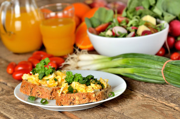 Wholemeal bread with scrambled eggs, fresh herbs, spring onions, tomatoes orange juice and salad bowl in background - healthy breakfast