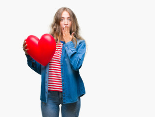 Fototapeta na wymiar Beautiful young blonde woman holding heart valentine over isolated background cover mouth with hand shocked with shame for mistake, expression of fear, scared in silence, secret concept