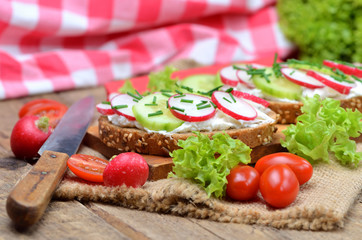 Close-up of grain bread with spread, fresh cucumber, radish, tomatoes and knife on a wooden cutting board - concept of healthy fitness breakfast or snack