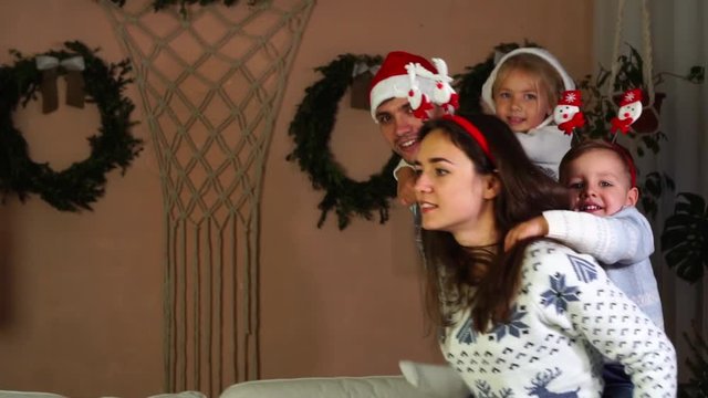 Cute family playing piggy back ride father and daughter chasing mother with son in Christmas sweaters funny Santa hats
