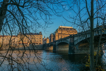 View of National theatre and buildings standing on riverbank, Prague, Czech Republic, granite bridge of Legions across Moldau river, branches and trees in foreground, fall evening, clear blue sky