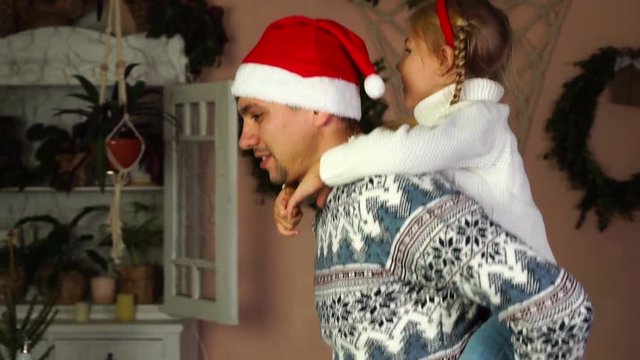 Excited dad in knitted Christmas sweater funny Santa Claus hat playing piggy back ride with cute daughter in slow motion