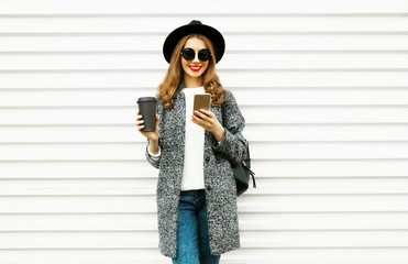 Fashion smiling woman using smartphone with coffee cup on white wall background