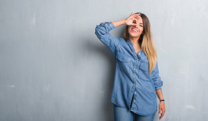 Young adult woman over grunge grey wall wearing denim outfit doing ok gesture with hand smiling, eye looking through fingers with happy face.