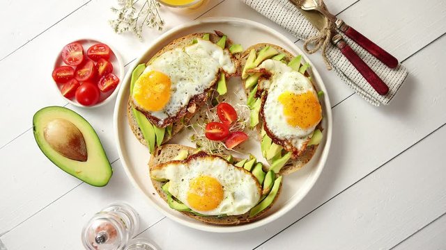 Delicious healthy breakfast with sliced avocado sandwiches with fried egg on top of bread. With orange juice, cherry tomatoes, radish sprouts, salt and peper. Flat lay, top view.