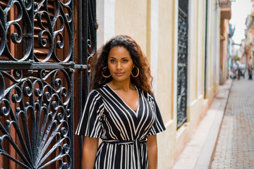 Young black lady with curly hair and a black and white dress posing in front of an old wooden door with an iron grid in Cuba Havana.