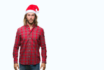 Young handsome man with long hair wearing santa claus hat over isolated background with serious expression on face. Simple and natural looking at the camera.