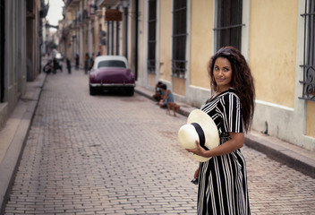 Young black woman with long curly hair standing in the streets of Havana in Cuba. She wears a black and white dress and has a typical cuban hat in her hand. 