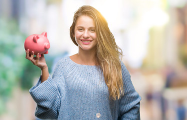 Young beautiful blonde woman holding piggy bank over isolated background with a happy face standing...