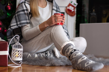 Girl sitting on furry carpet in home clothes and Christmas socks and warming with blanket. Woman holds cup of tea on leg and sitting near Xmas tree. Christmas and New Year concept