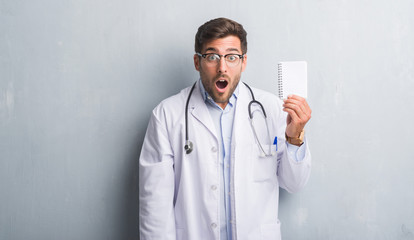 Handsome young doctor man over grey grunge wall holding blank notebook scared in shock with a surprise face, afraid and excited with fear expression