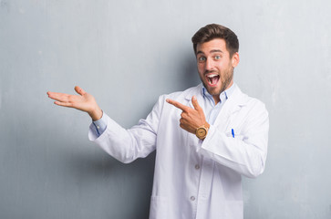 Handsome young professional man over grey grunge wall wearing white coat amazed and smiling to the camera while presenting with hand and pointing with finger.
