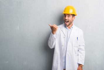 Handsome young engineer man over grey grunge wall wearing safety helmet pointing and showing with thumb up to the side with happy face smiling