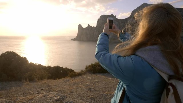 Young Attractive Woman Watching Sunset or Sunrise Over Bay from Top of the Mountain, Girl Takes Panorama Photos On Phone.Hair in the Wind. Handheld Atmospheric Shot. Crimea, Ukraine