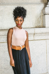 Portrait of Young African American Woman in New York. Young black female college student with afro hairstyle wearing sleeveless light color top, brown belt, black skirt, standing against vintage wall