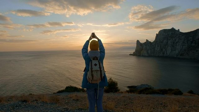 young attractive girl hiking makes photo on smartphone. Shoots a beautiful sunset on a cape by the sea. Reached the goal. Crimea, Ukraine. The concept of freedom and unity with nature.