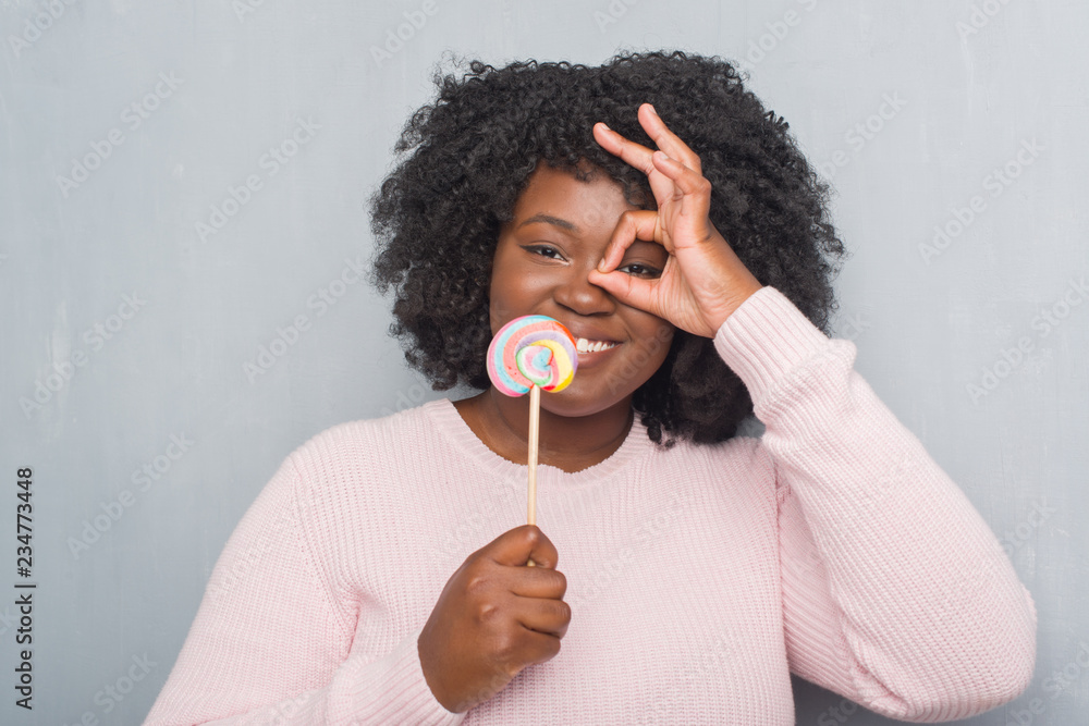 Canvas Prints Young african american woman over grey grunge wall eating lollipop candy with happy face smiling doing ok sign with hand on eye looking through fingers