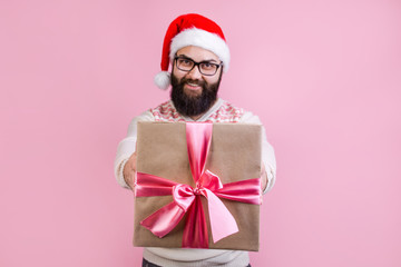 Happy bearded man in Santa Claus hat holds Christmas gift box in front of him standing on pink background