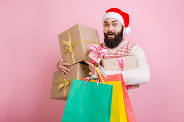 Happy man in Santa Claus hat stands on pink background with packages and gift boxes for Christmas
