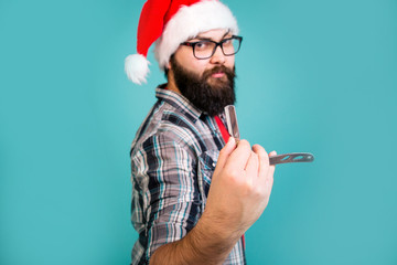 Bearded man in Santa Claus hat shows a dangerous razor stretching his hand forward to the foreground standing on a blue background