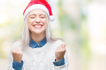 Young blonde woman wearing christmas hat over isolated background very happy and excited doing winner gesture with arms raised, smiling and screaming for success. Celebration concept.