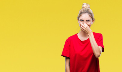 Young beautiful blonde woman wearing red t-shirt and glasses over isolated background smelling something stinky and disgusting, intolerable smell, holding breath with fingers on nose