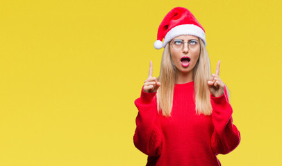 Fototapeta na wymiar Young beautiful blonde woman wearing christmas hat over isolated background amazed and surprised looking up and pointing with fingers and raised arms.