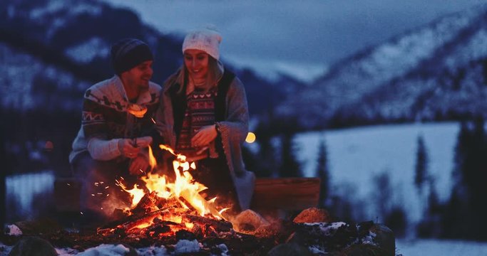 Couple Sitting by the Burning Campfire With Winter Mountains in Background. SLOW MOTION. Fairytale snow covered view with fire burning outdoors at dusk. 