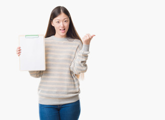 Young Chinese woman over isolated background holding clipboard pointing and showing with thumb up to the side with happy face smiling