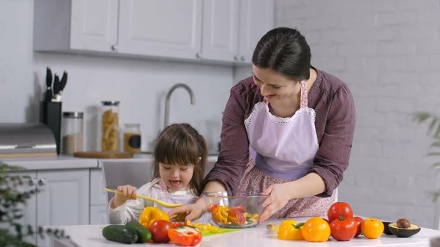 Cute little daughter with down syndrome helping her loving mother to prepare healthy fresh salad in domestic kitchen. Positive child with special needs and mother cooking healthy food in the kitchen.