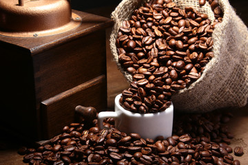 Fresh roasted coffee beans in burlap sack, coffee cup and grinder on dark background.