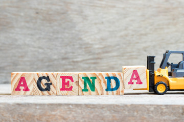 Toy forklift hold letter block a to complete word agenda on wood background