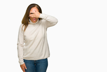Beautiful middle age adult woman wearing winter sweater over isolated background peeking in shock covering face and eyes with hand, looking through fingers with embarrassed expression.