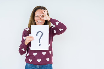Fototapeta premium Middle age woman thinking and holding paper with question mark symbol over isolated background with happy face smiling doing ok sign with hand on eye looking through fingers