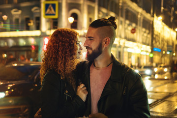 Couple in love on date. Boyfriend hugs girlfriend. She is holding an electronic cigarette. They walk on streets of night city. Wet asphalt after rain.