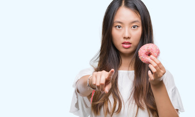 Young asian woman eating donut over isolated background pointing with finger to the camera and to you, hand sign, positive and confident gesture from the front