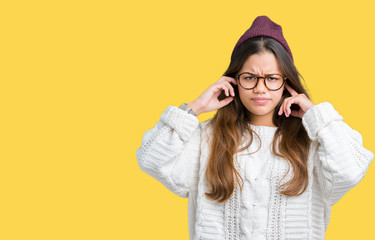 Young beautiful brunette hipster woman wearing glasses and winter hat over isolated background covering ears with fingers with annoyed expression for the noise of loud music. Deaf concept.