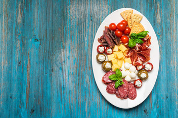 Various appetizers of cold meat, cheese, bacon, crispy cracker, olives are served on a white dish on a blue wooden background. Antipasto is a traditional Italian. Copy space.  Flat layout.