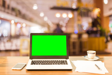Mockup image of laptop with blank green screen on wooden table blurred images In the coffee shop at shopping mall background.