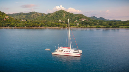 A nice drone shot of a large white catamaran yacht anchoring in a beautiful blue bay in front of an...