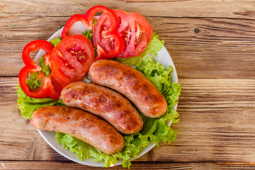 Ceramic plate with grilled sausages, sliced tomatoes and lettuce leaves on wooden table. Top view