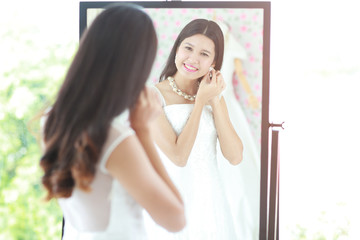 Portrait of beautiful Asian bride put on earring looking in mirror, her back out of focus large copy space, horizontal layout and warm light